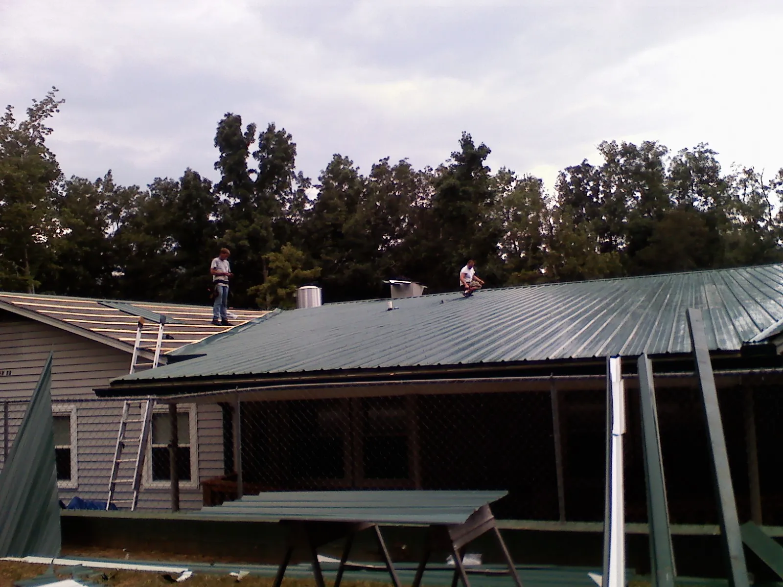 Two roofers installing green metal panels above a covered porch in Wildwood